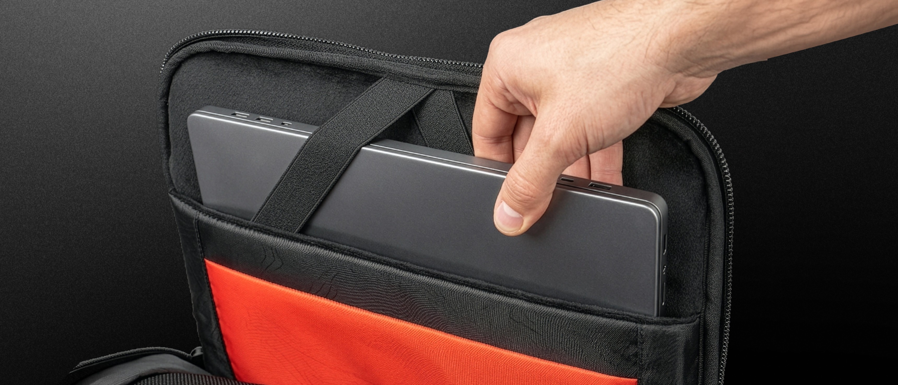 A hand placing a laptop into a padded compartment of a black and red bag.