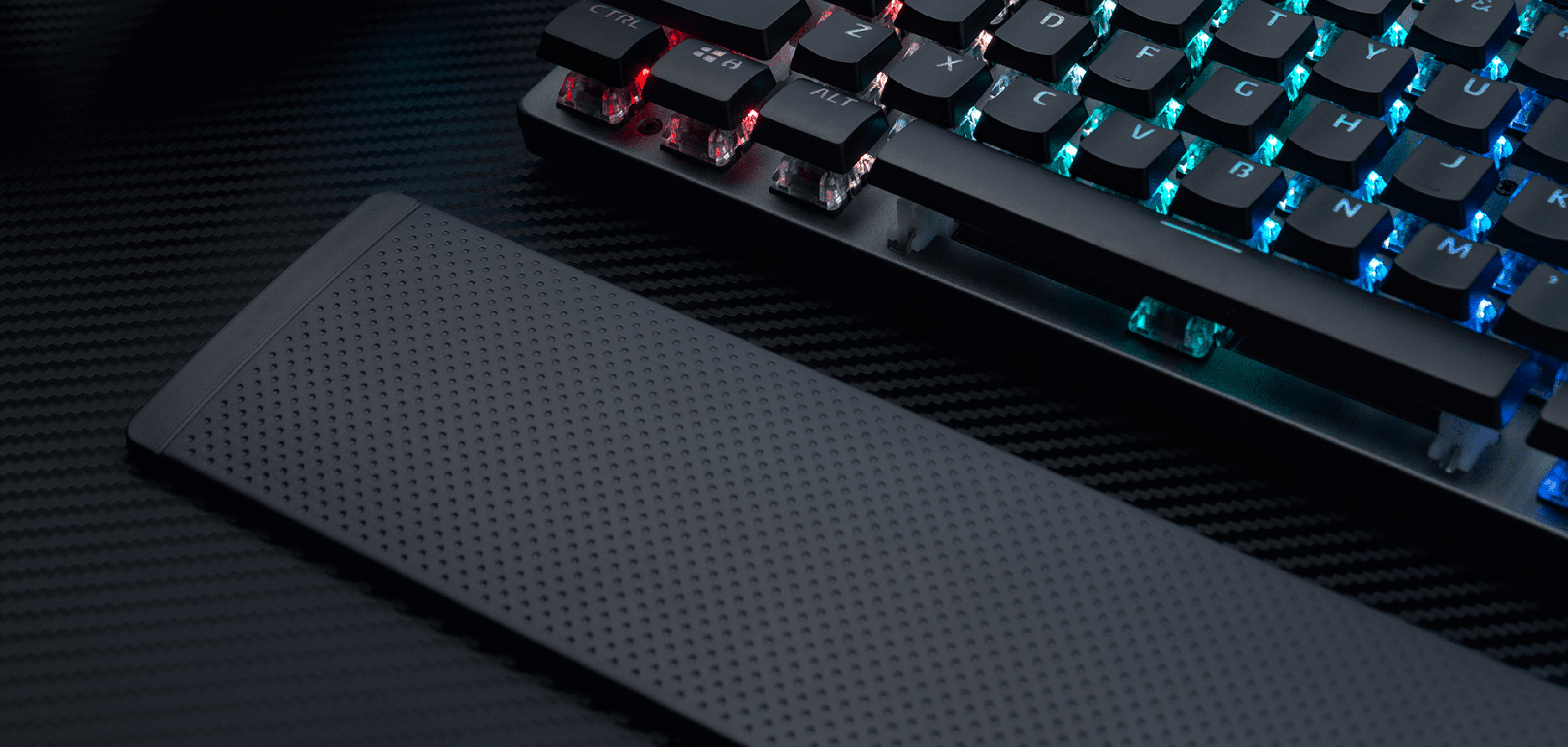 Gaming keyboard with illuminated keys and a textured wrist rest beside it on a dark surface.