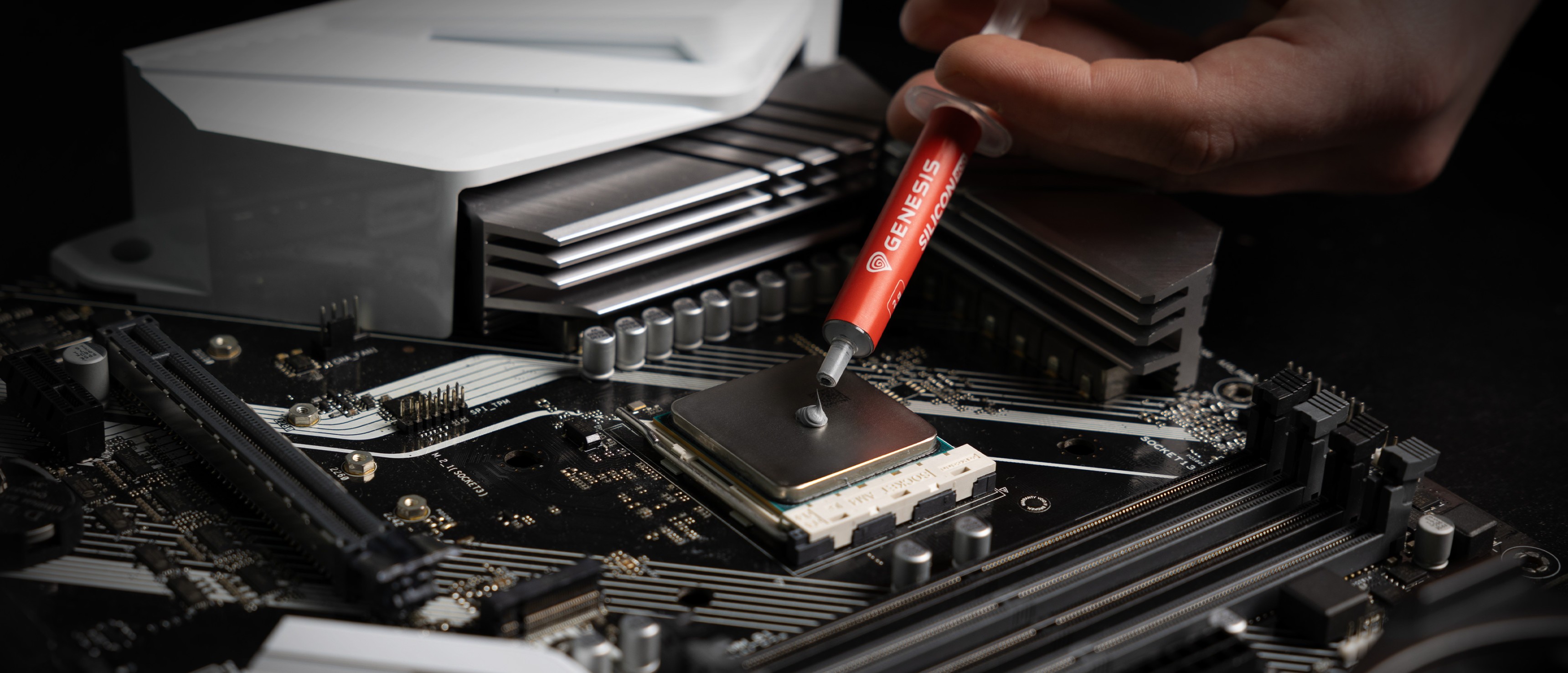 Thermal paste being applied from a syringe onto a CPU socket on a motherboard, with cooling components visible