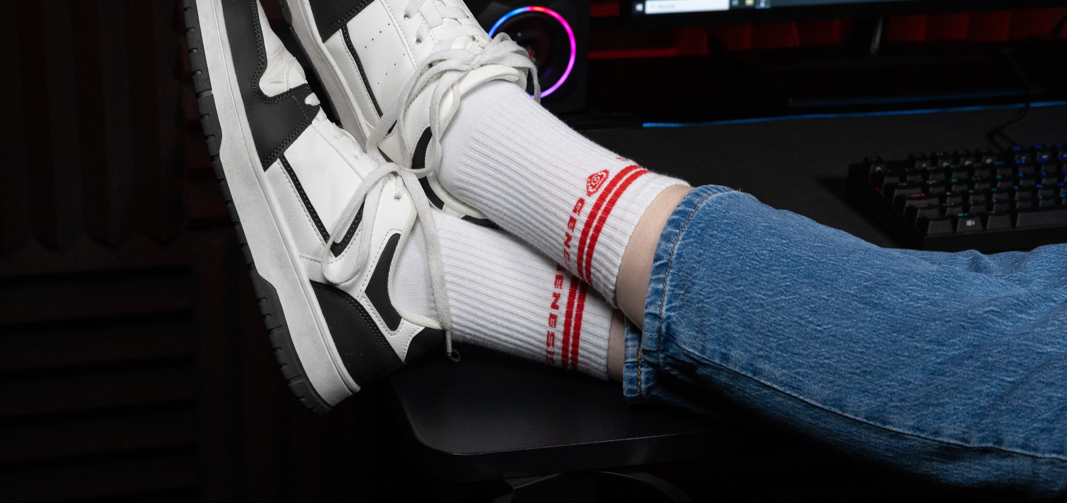 White sports socks with red branding, worn with white and black sneakers, resting on a desk with a computer setup