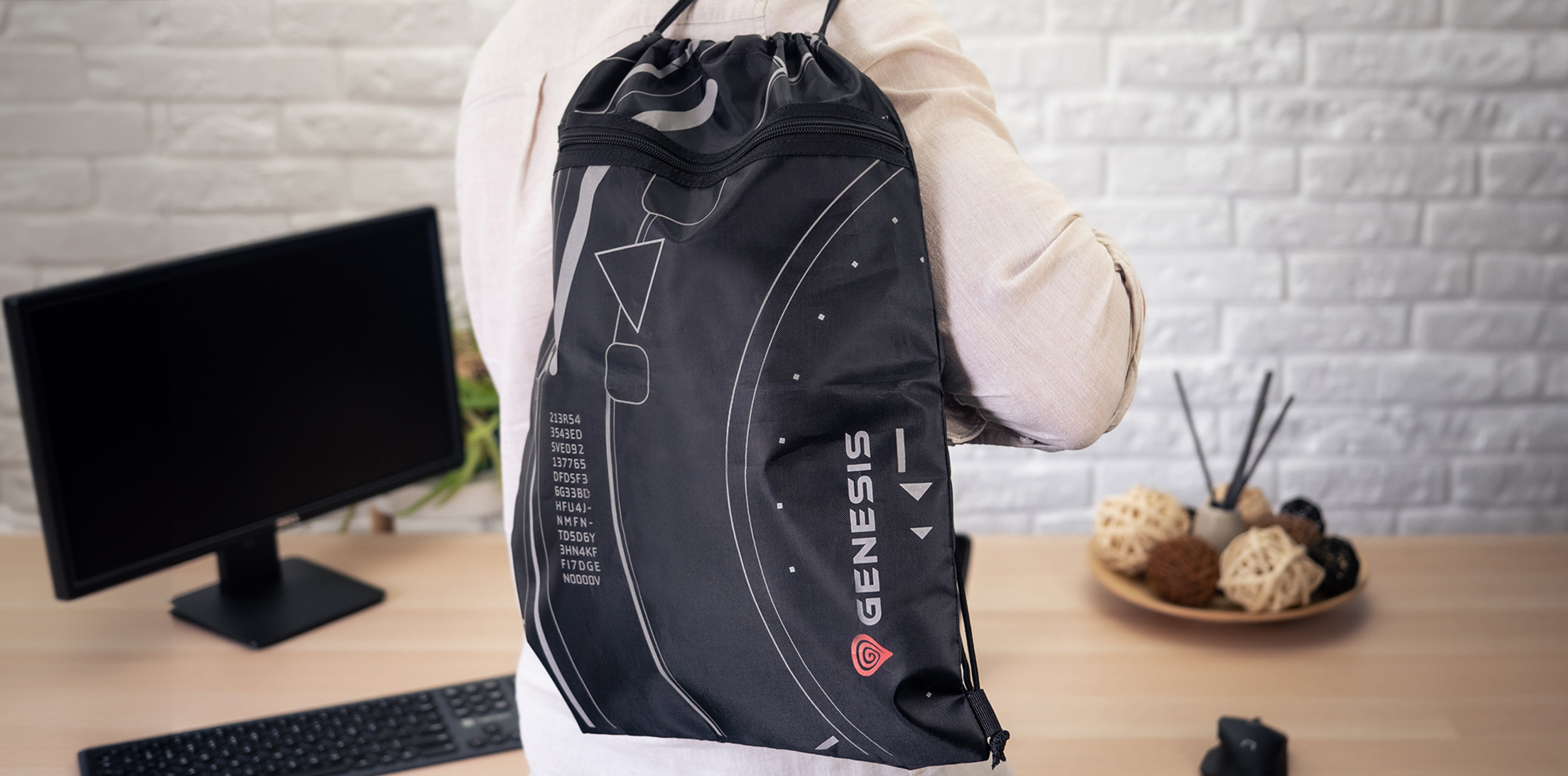 Drawstring backpack in black with white graphics, worn on the back of a person, set against a desk with a computer