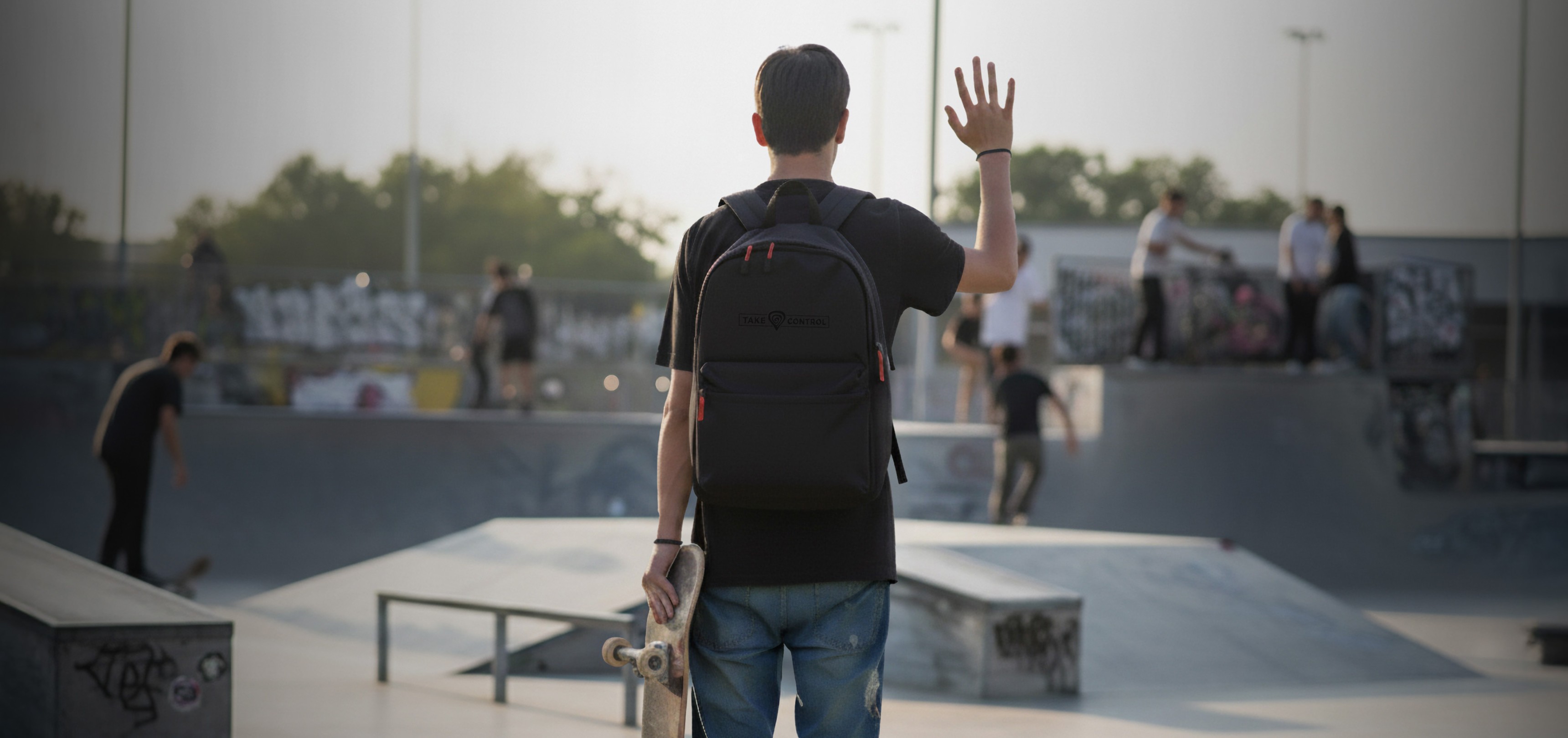 A young person at a skate park, wearing a black backpack, waves while holding a skateboard. The background features skaters and graffiti